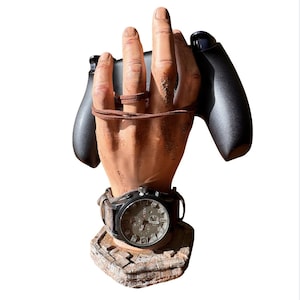 Puede incluir: Una escultura decorativa en forma de mano que sostiene un mando de videojuegos negro. La escultura presenta un reloj de pulsera con caja negra y correa de cuero marrón. La esfera del reloj es plateada con detalles en negro. La base es una estructura de piedra.