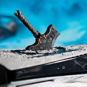 Puede incluir: Un hacha negra y dorada, con un mango detallado, está incrustada en una superficie gris, apoyada en una consola negra. El fondo es azul y blanco borroso. El hacha tiene intrincados detalles dorados.