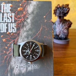 K&ouml;nnte beinhalten: Eine silberfarbene Uhr mit schwarzem Zifferblatt und gr&uuml;nem Nylonarmband liegt auf einem Buchcover. Das Zifferblatt der Uhr zeigt wei&szlig;e Ziffern und Zeiger. Das Buchcover tr&auml;gt den Titel "The Last of Us" und den Text "Joel Watch".