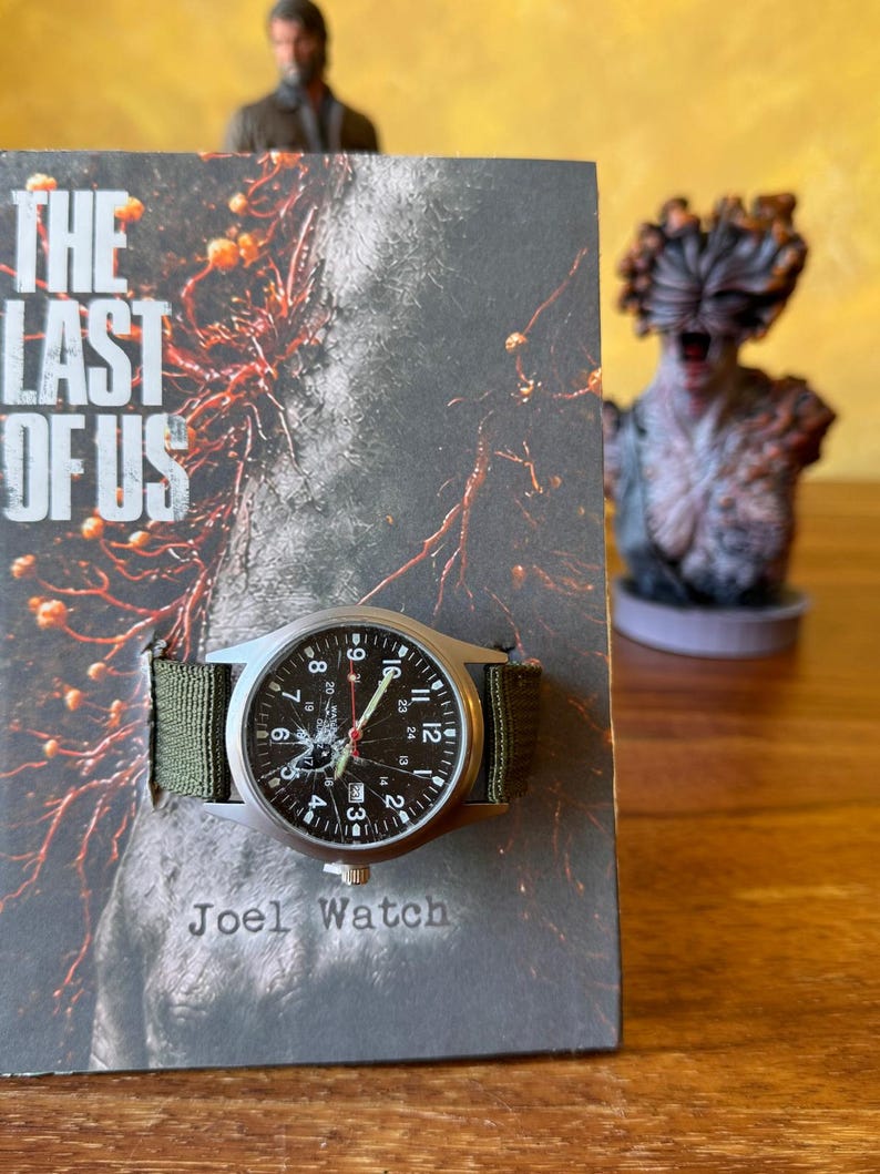K&ouml;nnte beinhalten: Eine silberfarbene Uhr mit schwarzem Zifferblatt und gr&uuml;nem Nylonarmband liegt auf einem Buchcover. Das Zifferblatt der Uhr zeigt wei&szlig;e Ziffern und Zeiger. Das Buchcover tr&auml;gt den Titel "The Last of Us" und den Text "Joel Watch".
