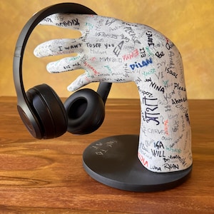 May include: A black wireless headphone set rests on a white hand-shaped stand. The stand is covered in handwritten names and phrases in various colors. The base of the stand is black and circular.