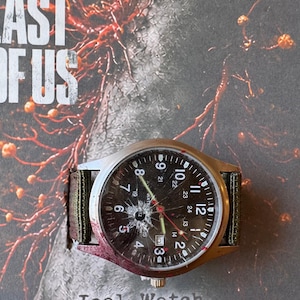 K&ouml;nnte beinhalten: Eine silberfarbene Armbanduhr mit zerbrochenem Zifferblatt und einem gr&uuml;nen und braunen Stoffarmband. Das schwarze Zifferblatt zeigt wei&szlig;e Ziffern und rote Zeiger. Der Text "Joel Watch" ist darunter zu sehen.