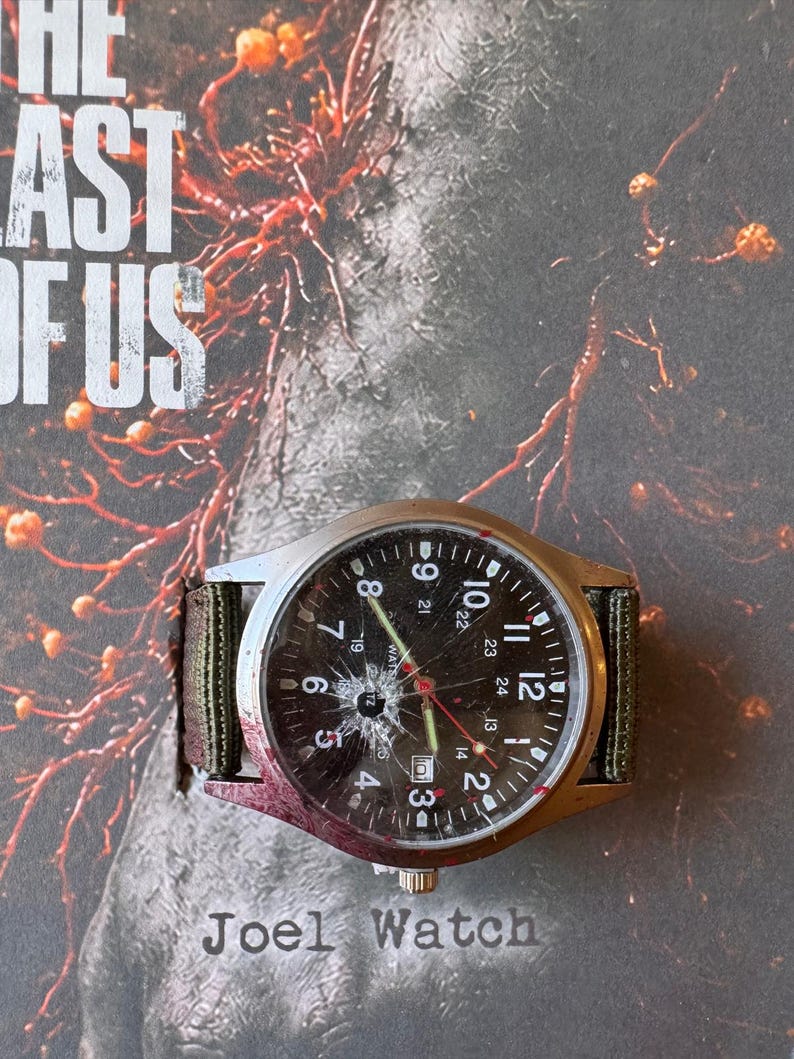 K&ouml;nnte beinhalten: Eine silberfarbene Armbanduhr mit zerbrochenem Zifferblatt und einem gr&uuml;nen und braunen Stoffarmband. Das schwarze Zifferblatt zeigt wei&szlig;e Ziffern und rote Zeiger. Der Text "Joel Watch" ist darunter zu sehen.
