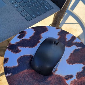 May include: A black wireless computer mouse on a round mousepad with a brown and white cowhide pattern.