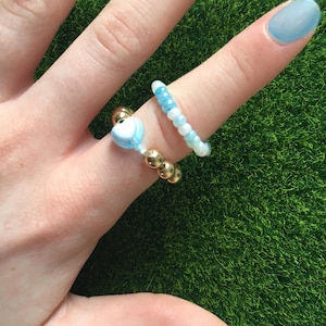 Op de afbeelding: Twee kralenringen op een vinger. Een ring is gemaakt van gouden kralen en heeft een blauwe hartbedel. De andere ring is gemaakt van blauwe en witte kralen.