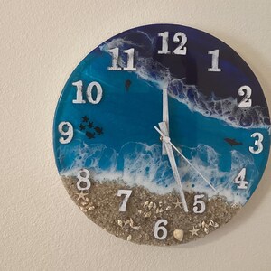 May include: Round wall clock with a beach-themed design. The clock face features a blue and white ocean scene with white numerals. The bottom of the clock has a sand-like texture with small seashells. The clock hands are silver.