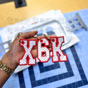May include: Three white fabric letters with red polka dots and red stitched borders, specifically the letters X, 6, and K, are attached to gold-toned metal key rings. The letters feature a block-style font and are held together as a set.