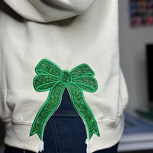 Może przedstawiać: Jas sportowy w jasnobeżowym kolorze z zieloną błyszczącą kokardką wszytą z tyłu. Kokardka jest umieszczona tak, aby wyglądało, jakby wystawała z tyłu bluzy.