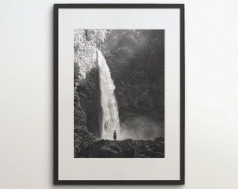 May include: A black and white photograph of a waterfall in a lush forest. A person stands at the base of the waterfall, looking up at the cascading water.