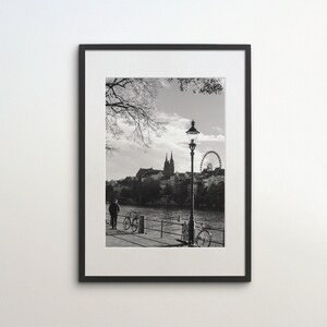 May include: Black and white photograph of a city skyline with a ferris wheel in the background. A street lamp stands in the foreground with two bicycles parked nearby. A person walks along the sidewalk.