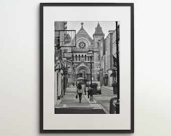 May include: Black and white photograph of a narrow street in a city. A church with a clock tower is visible at the end of the street. A person is walking down the street.