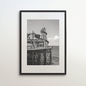 Könnte beinhalten: Schwarz-Weiß-Druck eines gerahmten Bildes eines Piers, der sich ins Meer erstreckt. Der Pier verfügt über ein Karussell, einen Turm mit Spiralmuster und einen Restaurantbereich. Das Bild ist eine Graustufenfotografie.