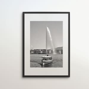 Może przedstawiać: Czarno-białe zdjęcie łodzi na jeziorze z dużą fontanną w tle. Fontanna to wysoki strumień wody strzelający wysoko w powietrze.