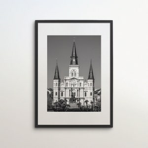 Puede incluir: Una fotografía en blanco y negro de la Catedral de San Luis en Nueva Orleans, Luisiana. La catedral es un edificio grande y ornamentado con dos torres altas y una cúpula central. La fotografía está enmarcada en un marco negro.
