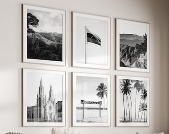 May include: A living room featuring a white sofa and a white ottoman. Six black and white framed photographs adorn the wall above the sofa, showcasing a mountain range, a flag, a church, and palm trees. A coffee table sits in front of the sofa, adorned with a vase of dried flowers and a book.