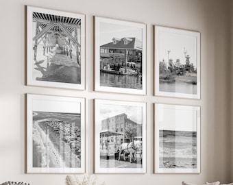 May include: A living room interior with a white sectional sofa, a white coffee table, and a white ottoman. The walls are white and there are six black and white framed photographs on the wall. The photographs are of various scenes, including a pier, a building, and a beach.