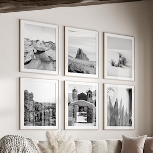 May include: A living room interior with a white sofa and a coffee table. The walls are decorated with six black and white framed photographs of desert landscapes and architecture. A large window with wooden trim looks out onto a green lawn.