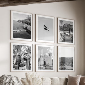May include: A living room with a white sofa and a white ottoman. There are six black and white framed photos on the wall above the sofa. The photos feature a beach scene, a flag, a boat, a church, and a mountain range. There is a large window with a view of trees and a cactus.