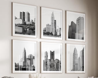 May include: A living room interior with a white sectional sofa, a round coffee table, and a white ottoman. Six black and white framed photographs of cityscapes are displayed on the wall above the sofa. The room has a large window with a view of trees and a wooden floor.