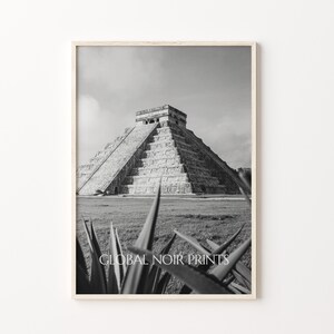 May include: Black and white photograph of the Mayan pyramid, Chichen Itza, in Mexico. The pyramid is made of stone and has a series of steps leading to the top. The pyramid is surrounded by grass and trees.