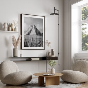 May include: A living room interior design with two plush armchairs, a coffee table, a black shelf with books, a black wall lamp, and a black and white photograph of a Mayan pyramid.