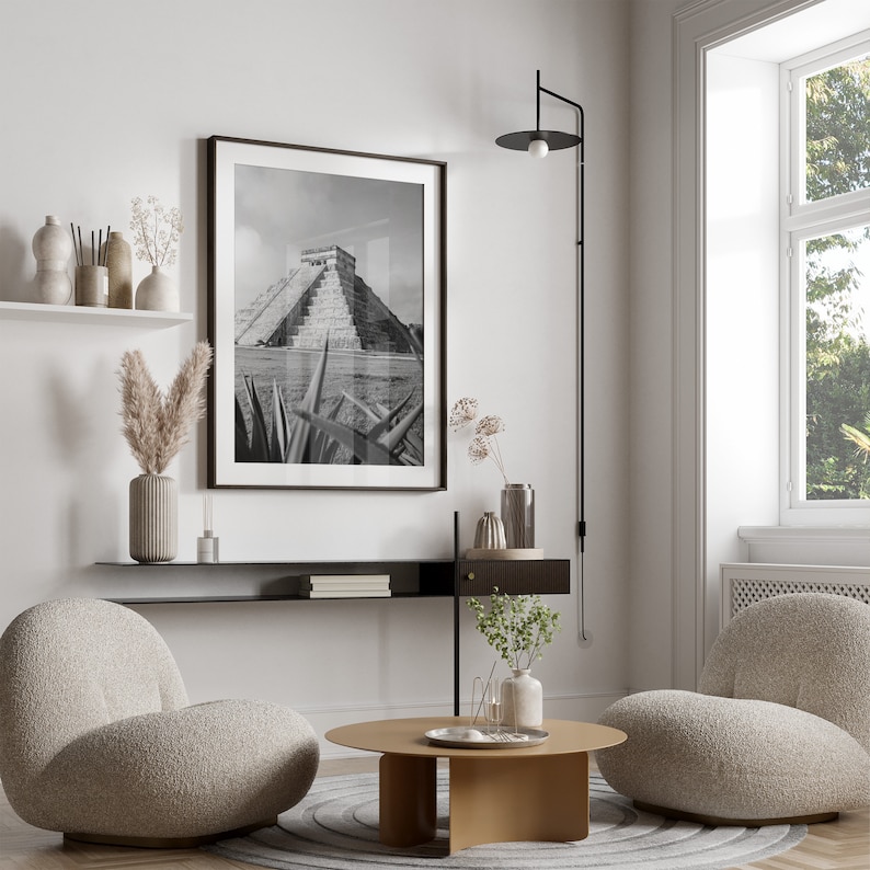 May include: A living room interior design with two plush armchairs, a coffee table, a black shelf with books, a black wall lamp, and a black and white photograph of a Mayan pyramid.