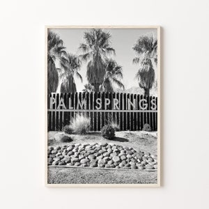 Może przedstawiać: Czarno-białe zdjęcie znaku z napisem "PALM SPRINGS" białymi literami. Znak znajduje się za drewnianym płotem z palmami w tle.