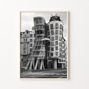 May include: Monochrome photograph of a modern building with a distinctive, curved glass facade. The building features a tower-like structure and a wavy design. The image is framed in a light wood frame, suitable for home decor.