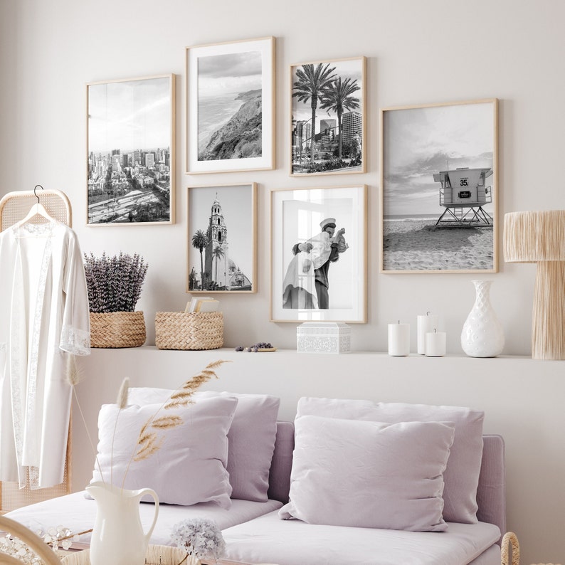 Op de afbeelding: Een galeriewand met zwart-witfoto's van een skyline, een strandscene, een stel en een gebouw. De foto's zijn ingelijst in zwarte houten frames en hangen aan een witte muur.