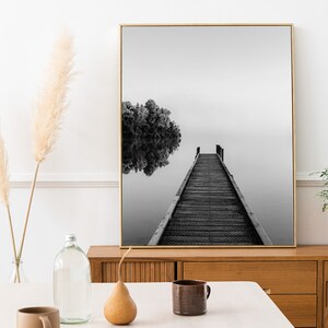 May include: Black and white photograph of a wooden dock extending into a still lake. The dock leads to a small island with trees. The photo is framed in a gold frame and hanging on a white wall.