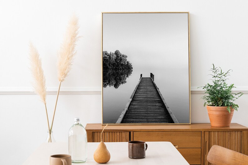 May include: Black and white photograph of a wooden dock extending into a still lake. The dock leads to a small island with trees. The photo is framed in a gold frame and hanging on a white wall.