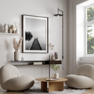 May include: A living room interior design with two plush armchairs, a coffee table, a black shelf with books, a black and white photograph of a wooden pier extending into a foggy lake, and a black wall sconce with a round light fixture.