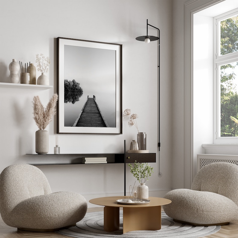 May include: A living room interior design with two plush armchairs, a coffee table, a black shelf with books, a black and white photograph of a wooden pier extending into a foggy lake, and a black wall sconce with a round light fixture.