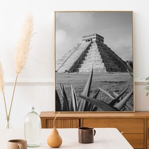 May include: Black and white photograph of the Mayan pyramid of Chichen Itza in Mexico. The pyramid is surrounded by grass and a few agave plants are in the foreground.