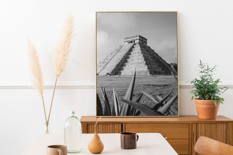 May include: Black and white photograph of the Mayan pyramid of Chichen Itza in Mexico. The pyramid is surrounded by grass and a few agave plants are in the foreground.