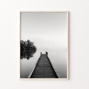 May include: A black and white photograph of a wooden dock extending into a misty lake. The dock is made of wooden planks and has a railing on either side. The lake is calm and still, and the mist is rising from the water. A small island with trees is visible in the background.