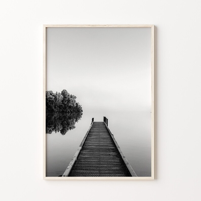 May include: A black and white photograph of a wooden dock extending into a misty lake. The dock is made of wooden planks and has a railing on either side. The lake is calm and still, and the mist is rising from the water. A small island with trees is visible in the background.