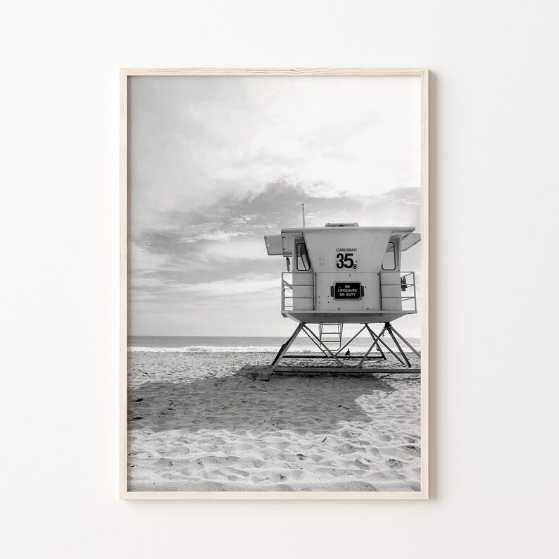 Op de afbeelding: Zwart-witfoto van een reddingsbrigadepost op een zandstrand. De post heeft een bord met de tekst "Carlsbad 35" en "Geen badmeester aanwezig".
