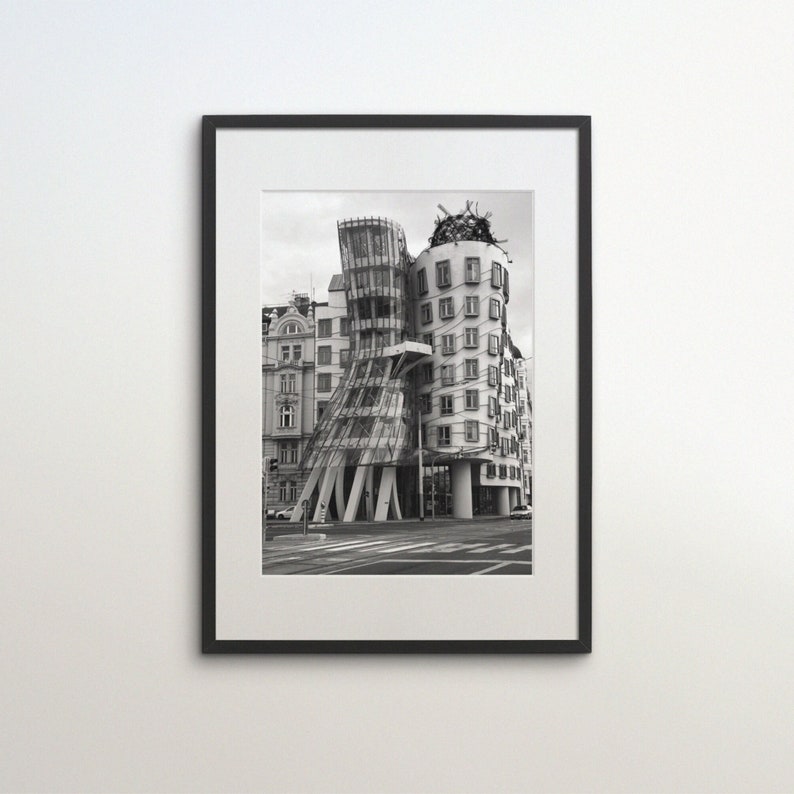 May include: Framed monochrome print of the Dancing House in Prague. The building's distinctive design includes curved glass and concrete features. The print is displayed on a white wall, with a black frame.
