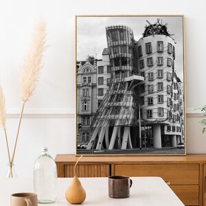 May include: Monochrome framed print of the Dancing House in Prague. The building has a distinctive, deconstructivist style with glass and concrete. The print is displayed on a wooden cabinet. Decorative items, including a plant and vase, are in the foreground.
