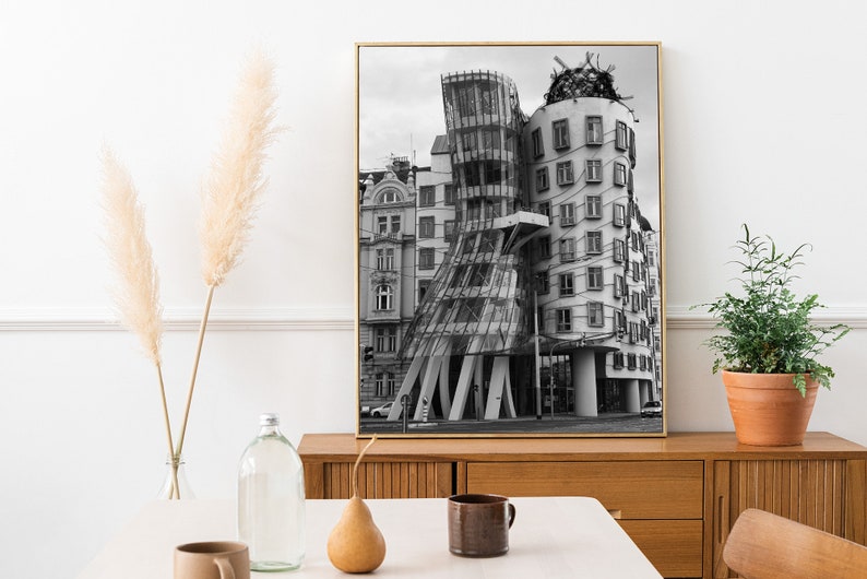 May include: Monochrome framed print of the Dancing House in Prague. The building has a distinctive, deconstructivist style with glass and concrete. The print is displayed on a wooden cabinet. Decorative items, including a plant and vase, are in the foreground.