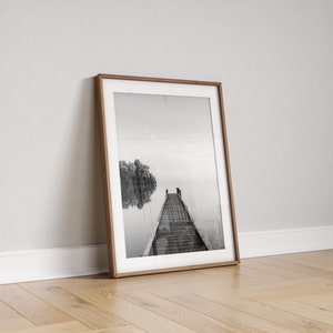 May include: Black and white photograph of a wooden dock extending into a misty lake. A small tree is reflected in the water.