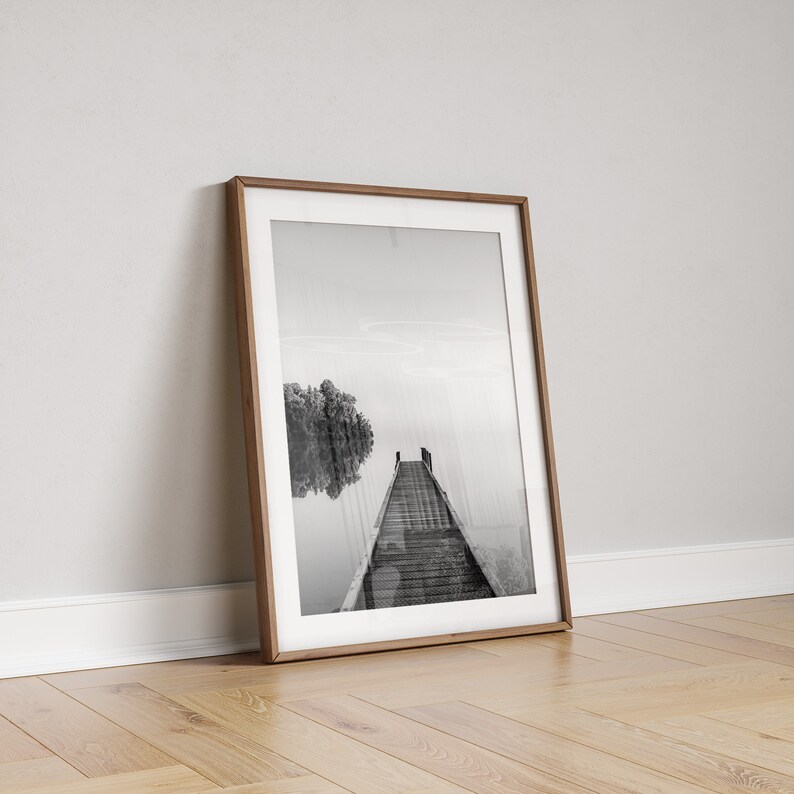 May include: Black and white photograph of a wooden dock extending into a misty lake. A small tree is reflected in the water.