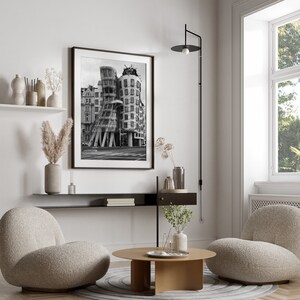May include: A contemporary living room with a framed monochrome photograph of a building. The room includes two beige armchairs, a round wooden coffee table, and a black wall-mounted lamp. Decorative vases and dried plants enhance the room's design.
