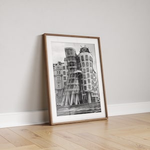 May include: Monochrome framed print of a distinctive, modern building with a curved facade. The print is in a brown wooden frame, leaning against a white wall and resting on a wooden floor.