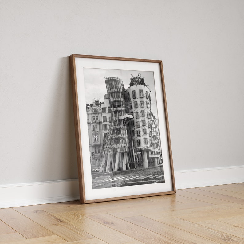 May include: Monochrome framed print of a distinctive, modern building with a curved facade. The print is in a brown wooden frame, leaning against a white wall and resting on a wooden floor.