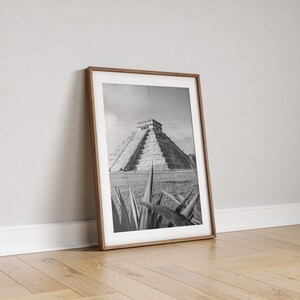 May include: Black and white photograph of the Mayan pyramid of Chichen Itza in Mexico, framed in a wooden frame. The pyramid is seen from a distance, with a foreground of agave plants.