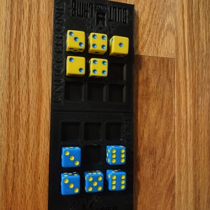 May include: A black dice tray with the words "Cult of the Lamb" printed on it. The tray has two rows of five compartments, each holding a yellow and blue dice.
