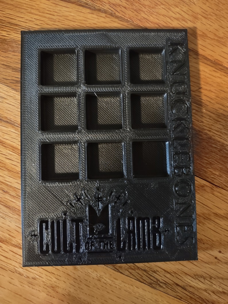 May include: Black plastic storage box with 12 square compartments. The box has the words "KNUCKLEBONES" printed vertically on the right side and "CULT OF THE CROWN" printed on the bottom.