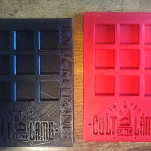 May include: Two 3D printed game trays, one black and one red, with a grid of nine squares. Both trays feature the text "Cult of the Lamb" and "Knick Knack Bones".